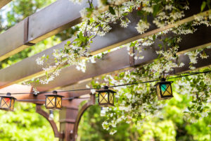 Patio outdoor spring garden in backyard of home with lantern lamps lights hanging from pergola canopy wooden gazebo and plants white flowers